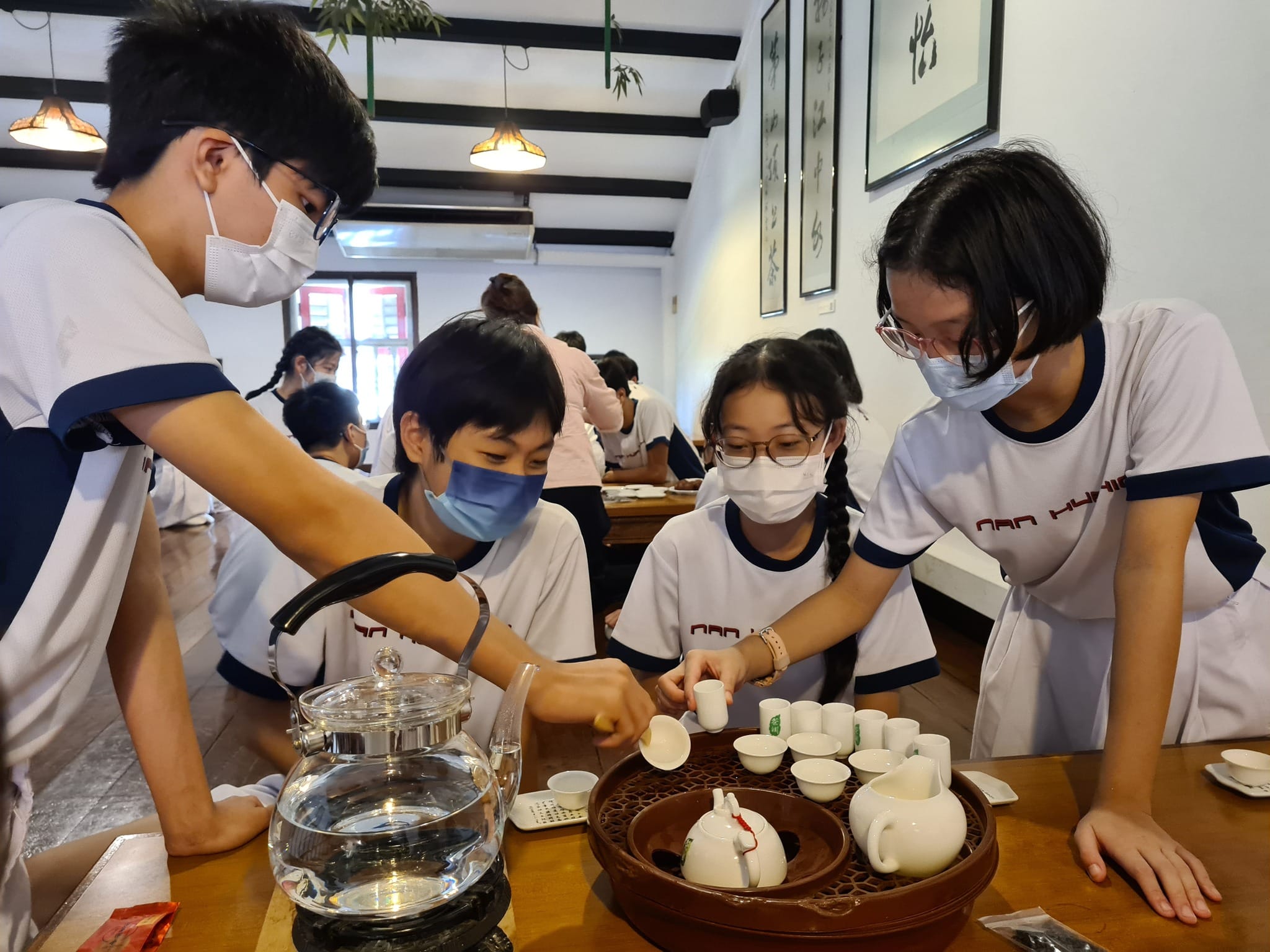 Four masked students in white uniforms gather around a wooden table with a Chinese tea set and glass kettle.